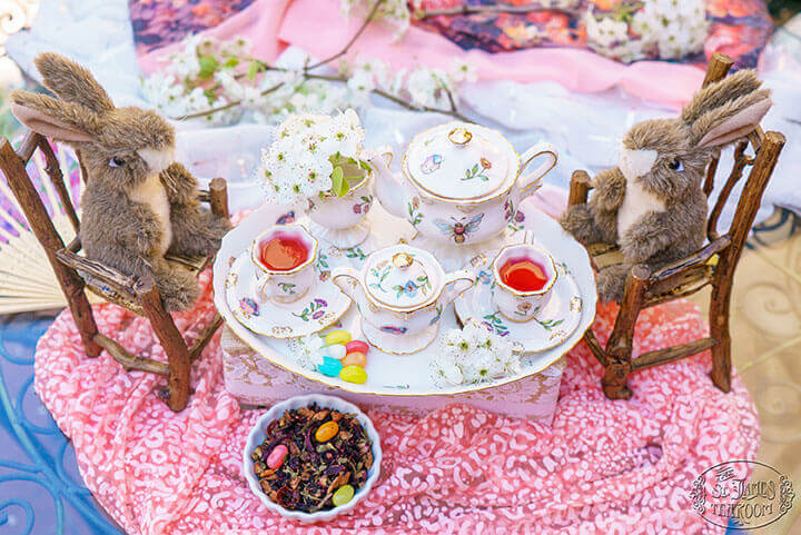 Two plush toy rabbits sitting on miniature wooden chairs at a small afternoon tea set table