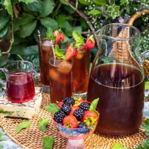 A wicker tray holds a pitcher and glasses of Iced Strawberry Tea garnished with mint and strawberries, a small glass pitcher of red liquid, and a bowl of mixed berries, all set outdoors among green plants.