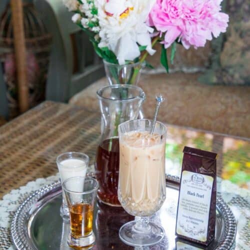 A silver tray holds a glass of iced coffee, a shot of cream, a shot of whiskey, a carafe, a bag of Silky Iced Tea, and a spoon. Pink and white flowers in a vase sit on the wicker table in the background.