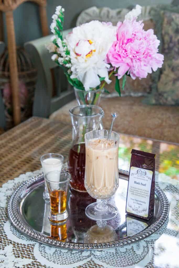 A silver tray holds a glass of iced coffee, a shot of cream, a shot of whiskey, a carafe, a bag of Silky Iced Tea, and a spoon. Pink and white flowers in a vase sit on the wicker table in the background.