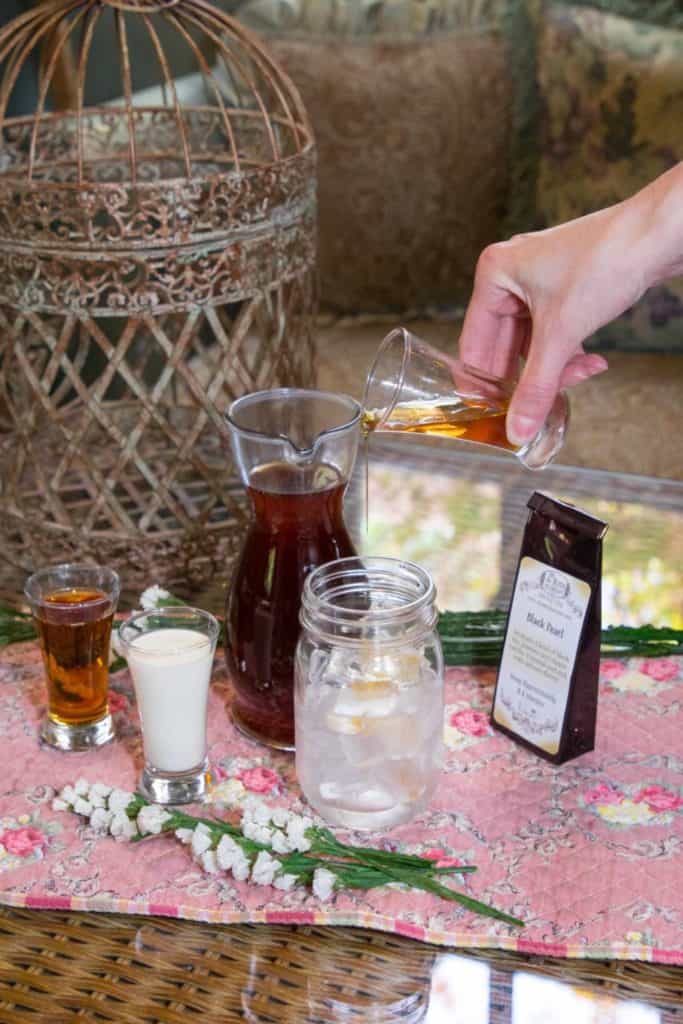 A hand pours Silky Iced Tea into a mason jar with ice, next to a pitcher of tea, a packet of black pearl tea, cream, and flowers on a floral tablecloth. A decorative birdcage is in the background.