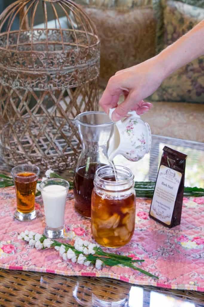 A hand pours cream from a floral pitcher into a mason jar of silky iced tea on a pink mat, with a small liquor shot, cream shot, coffee bag, carafe, and white flowers nearby.
