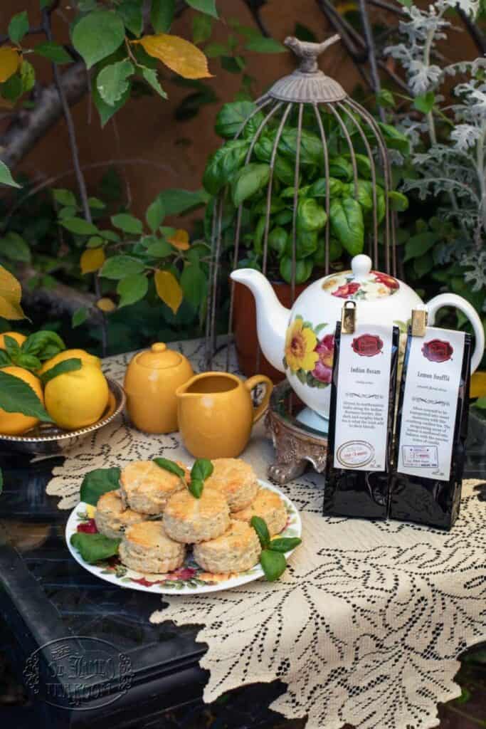 A floral teapot, golden cream and sugar jars, lemons, two tea packets, and a plate of basil lemon scones sit on a lace cloth-covered table in a garden with green leaves and yellow fruit.