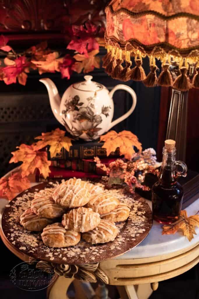 Vintage setting with a teapot, books, and maple leaves. A decorative plate holds glazed pastries inspired by an easy maple oat scone recipe, and a small bottle of syrup sits nearby under a patterned lampshade. Cozy autumn atmosphere.