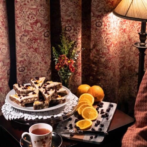A cozy scene with a plate of cake slices—perhaps an easy Thanksgiving gluten-free dessert recipe from The St. James Tearoom—a cup of tea, sliced oranges, and blueberries on a table beside a lamp, patterned curtains, and a small bouquet of flowers.
