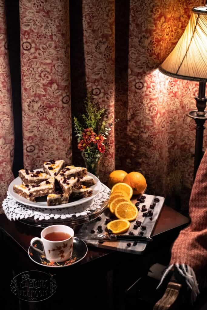 A cozy scene with a plate of cake slices—perhaps an easy Thanksgiving gluten-free dessert recipe from The St. James Tearoom—a cup of tea, sliced oranges, and blueberries on a table beside a lamp, patterned curtains, and a small bouquet of flowers.