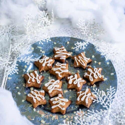 A blue plate with star-shaped cookies, reminiscent of a delicious gingerbread scone recipe from The St. James Tearoom, topped with white icing and surrounded by artificial snow and silver snowflake decorations, creating a festive winter scene.