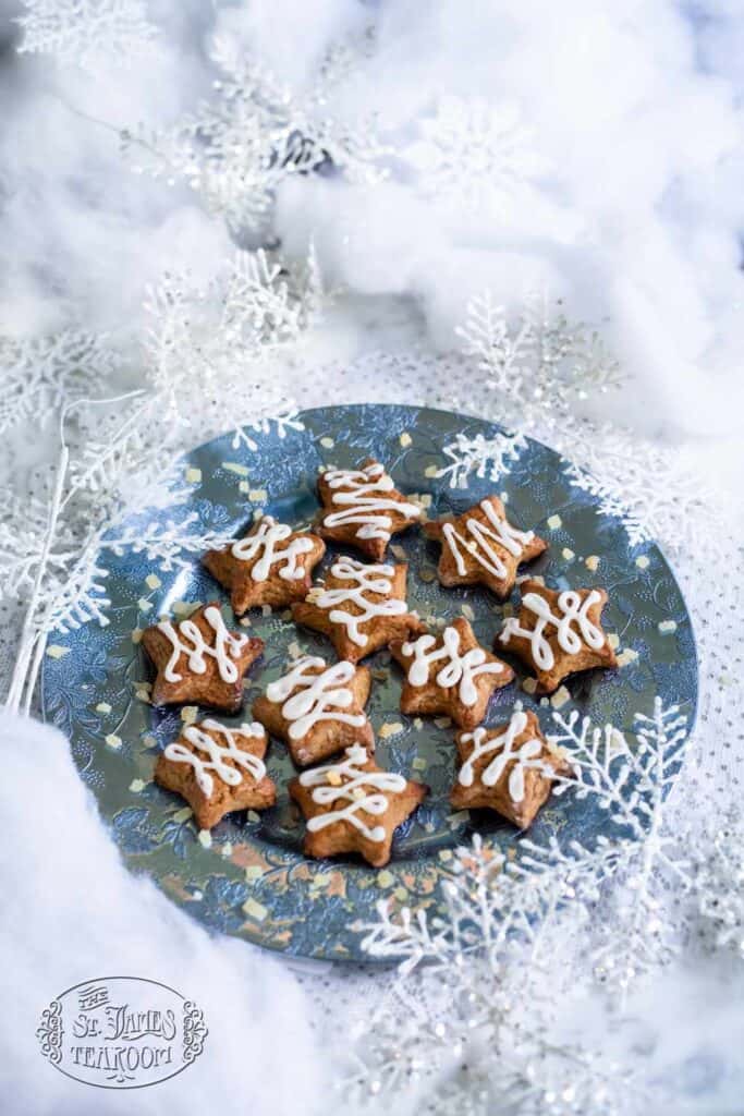 A blue plate with star-shaped cookies, reminiscent of a delicious gingerbread scone recipe from The St. James Tearoom, topped with white icing and surrounded by artificial snow and silver snowflake decorations, creating a festive winter scene.