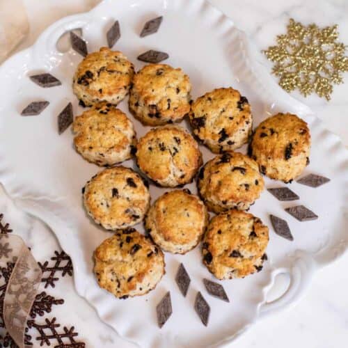 Ten golden-brown scones with chocolate chips are arranged neatly on a white decorative plate, surrounded by gold holiday ornaments and ribbons on a marble surface—a delightful treat inspired by our Quick and Easy Chocolate Cherry Scone Recipe from The St. James Tearoom.