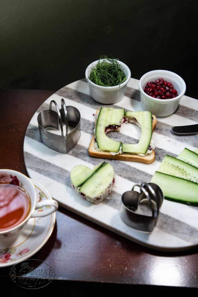 A striped tray holds a teacup, a slice of bread with cream and cucumber, metal sandwich cutters, cucumber slices, a bowl of berries, and a bowl of herbs—perfect accompaniments for enjoying the Quick and Easy Chocolate Cherry Scone Recipe from The St. James Tearoom.