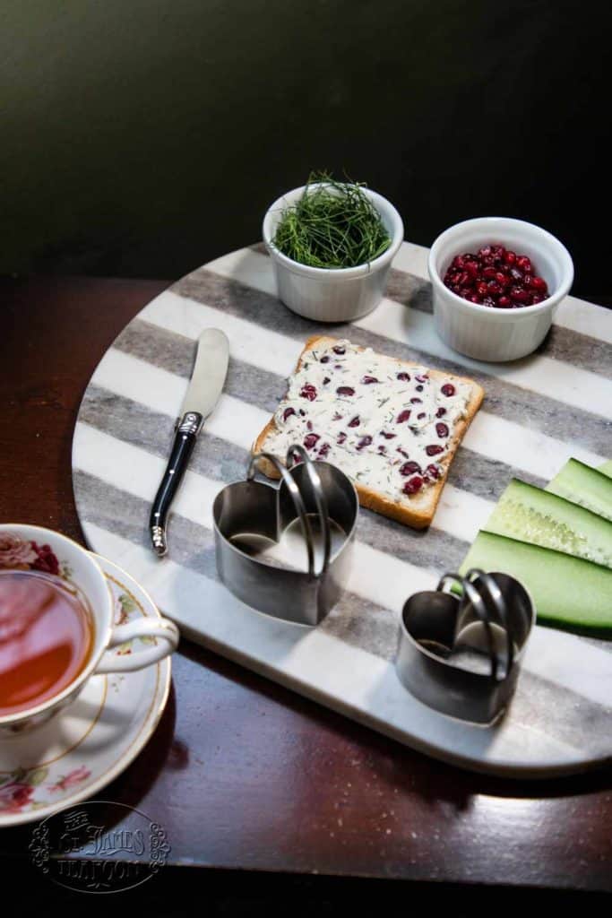 A striped tray displays a delightful array: a cracker topped with cream cheese and cranberries, a knife, cucumber slices, metal cookie cutters, chives, and a small bowl of pomegranate seeds. Perfectly paired with a cup of tea from The St. James Tearoom, this charming setup inspires you to try their Quick and Easy Chocolate Cherry Scone Recipe for your next gathering.