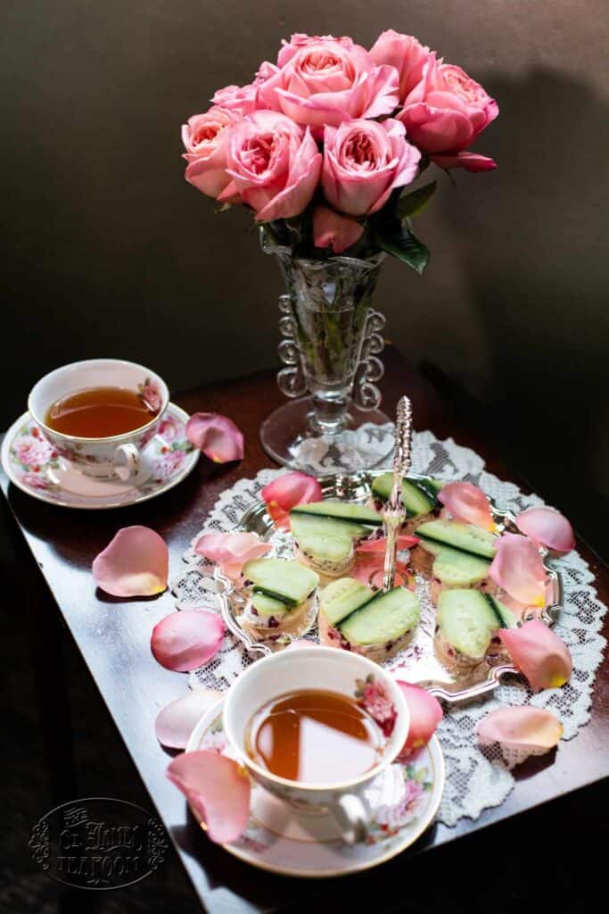 A small table with two teacups, a plate of cucumber sandwiches, pink rose petals, and a vase of pink roses, set for tea time on a lace doily—perfect for serving treats like the Quick and Easy Chocolate Cherry Scone Recipe from The St. James Tearoom.