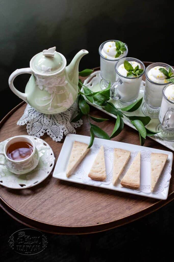 Enjoy a charming afternoon with a tea set featuring a teapot, a cup of tea, and the Best Traditional Shortbread Cookie Afternoon Tea Recipe from The St. James Tearoom. Indulge in delicate shortbread cookies on a plate alongside four glass cups of mousse topped with mint leaves, all beautifully arranged on a round wooden table with a lace doily.
