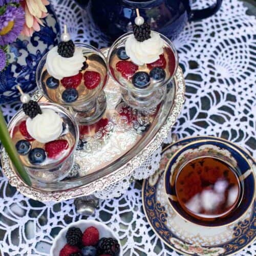 A silver tray holds parfaits topped with cream and berries, next to a cup of tea and a blue teapot on a lace tablecloth. A bowl of raspberries and blueberries sits nearby, accompanied by a bouquet of flowers—and for an extra touch of elegance, consider serving The St. James Tearoom’s Sparkling Wine Berry Gelatin Recipe alongside this delightful setting.