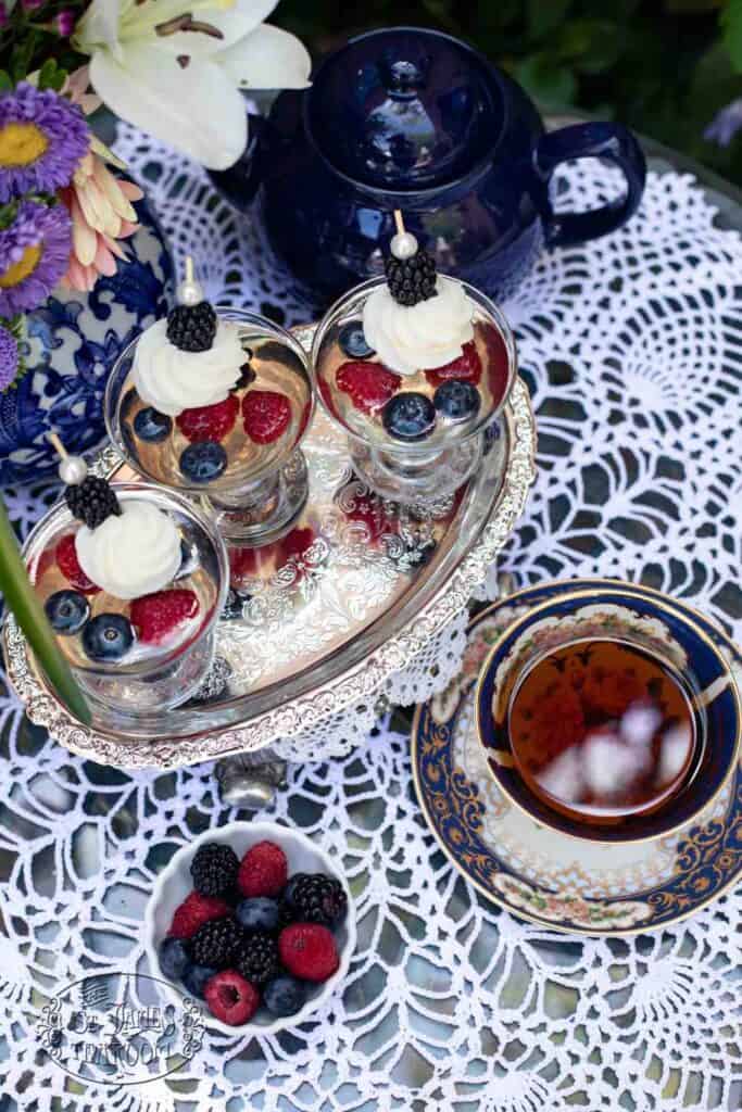 A silver tray holds parfaits topped with cream and berries, next to a cup of tea and a blue teapot on a lace tablecloth. A bowl of raspberries and blueberries sits nearby, accompanied by a bouquet of flowers—and for an extra touch of elegance, consider serving The St. James Tearoom’s Sparkling Wine Berry Gelatin Recipe alongside this delightful setting.