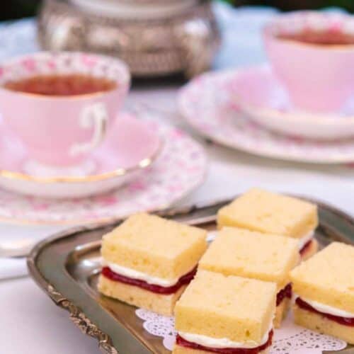 Six small Victoria Sandwich Cake sandwiches with cream and jam filling sit on a silver tray. In the background are pink teacups, saucers, and a teapot, all arranged on a white tablecloth for afternoon tea at The St. James Tearoom.