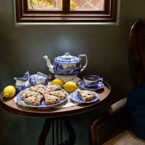 A wooden table with blue-and-white china teapot, teacups, cream and sugar set, lemon, and a plate of gluten-free blueberry almond scones sits by a window and an upholstered chair, with autumn leaves visible outside.