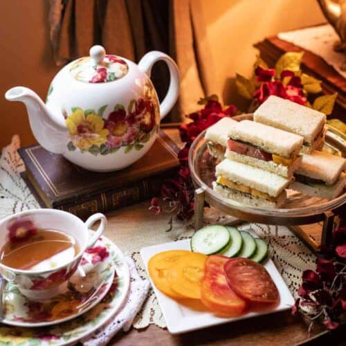 A floral teapot and teacup with tea, a plate of cucumber and tomato slices, and a tiered stand of assorted tea sandwiches—including a delightful Gluten-Free Herbed Tomato and Cucumber Tea Sandwich Recipe—are set on a cozy table with a doily and lamp.