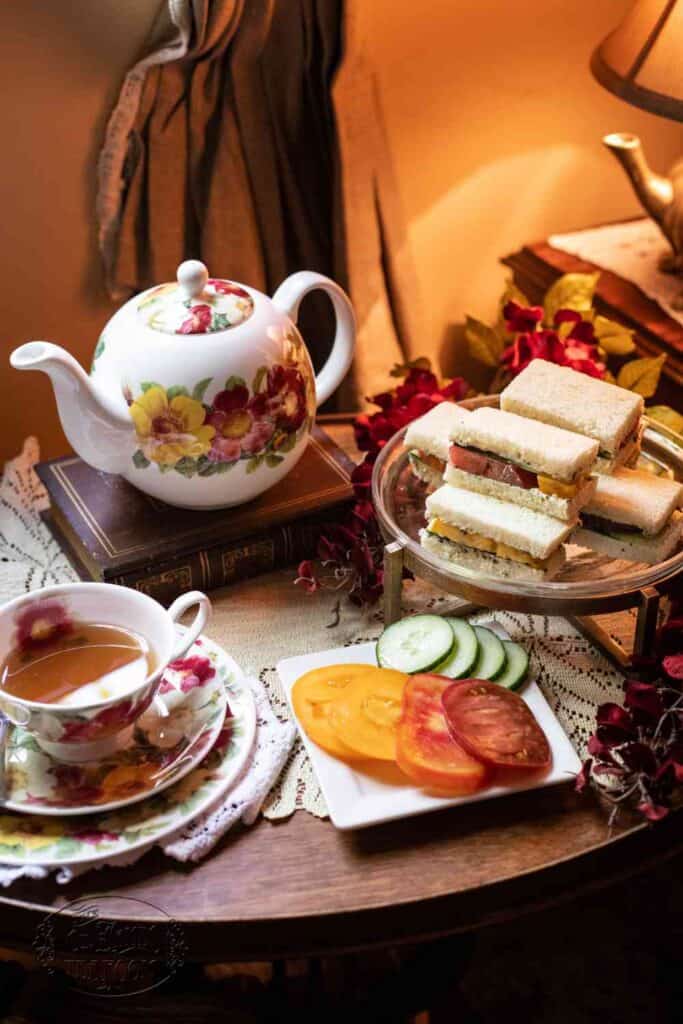 A floral teapot and teacup with tea, a plate of cucumber and tomato slices, and a tiered stand of assorted tea sandwiches—including a delightful Gluten-Free Herbed Tomato and Cucumber Tea Sandwich Recipe—are set on a cozy table with a doily and lamp.
