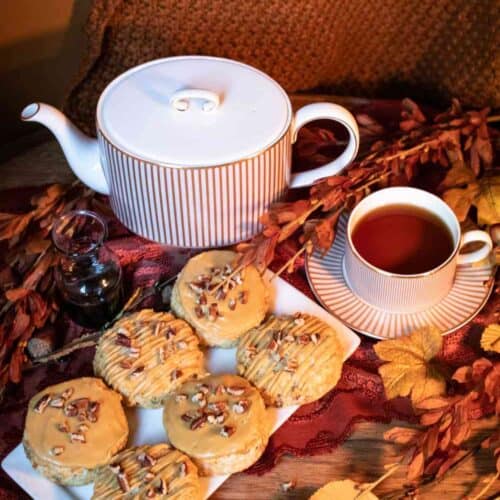 A teapot, cup of tea, and six iced pastries topped with chopped nuts—reminiscent of a Gluten-Free Pecan Scones Recipe with Molasses Glaze—sit on a wooden table decorated with autumn leaves and a red cloth, creating a cozy, fall-themed setting.