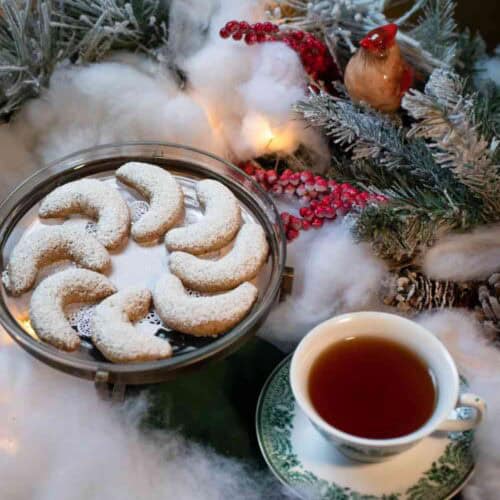 A plate of crescent cookies dusted with powdered sugar and a cup of tea sit on a table surrounded by snowy pine branches, red berries, and warm lights—setting the perfect scene for trying your favorite Christmas Cookies Recipe.