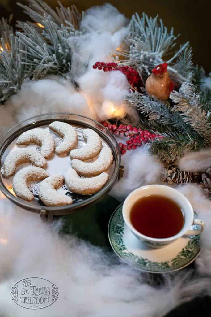 A plate of crescent cookies dusted with powdered sugar and a cup of tea sit on a table surrounded by snowy pine branches, red berries, and warm lights—setting the perfect scene for trying your favorite Christmas Cookies Recipe.