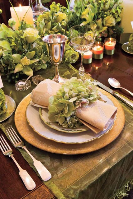 Elegant table setting with gold-rimmed plates, silverware, wine glasses, beige napkins with green floral accents, a green tablecloth, candles, and lush green floral centerpieces—this refined arrangement truly embodies the definition of hospitality.