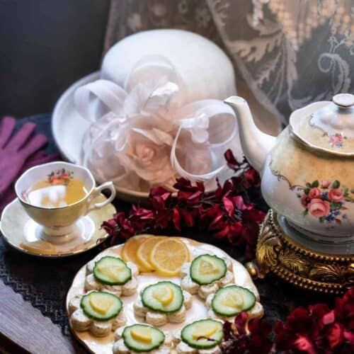 A vintage tea set with a teacup, saucer, and teapot sits beside cucumber sandwiches inspired by a classic Lemon Dill Cucumber Tea Sandwich Recipe, lemon slices, a white hat with flowers, red gloves, and lace curtains on a wooden table.