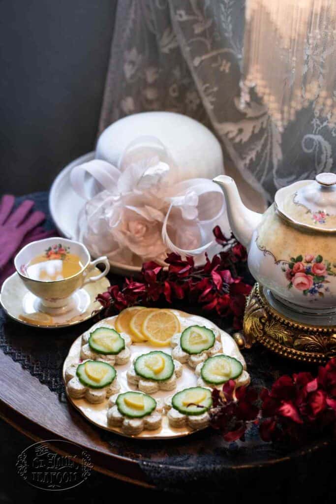 A vintage tea set with a teacup, saucer, and teapot sits beside cucumber sandwiches inspired by a classic Lemon Dill Cucumber Tea Sandwich Recipe, lemon slices, a white hat with flowers, red gloves, and lace curtains on a wooden table.