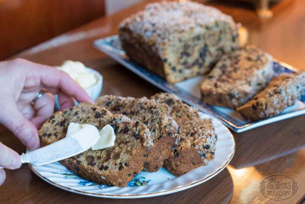 A hand spreads butter on a slice of fruit-studded bread with streusel topping, reminiscent of a classic Irish Tea Bread Recipe. More slices and a loaf sit on a plate and platter behind, on a wooden table.