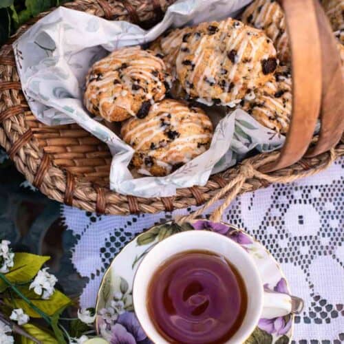 A basket of glazed scones with chocolate chips, inspired by a delightful Easter Scones Recipe, sits on a lace tablecloth beside a floral teacup filled with tea. Flowers and greenery are arranged around the setup.