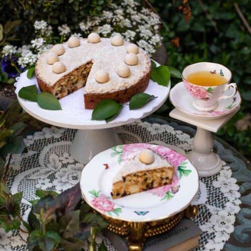 A beautifully decorated Simnel Cake, following a classic Simnel Cake Recipe for Mother’s Day, is topped with golden marzipan balls and sits on a white cake stand. A generous slice rests on a floral plate beside a teacup, saucer, and green leaves, all arranged atop a lace tablecloth outdoors.