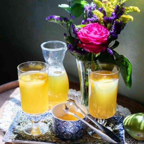 Two glasses and a carafe of yellow juice with ice sit on a tray with a bowl, spoon, and floral centerpiece with pink, yellow, and purple flowers—all arranged on a lace doily in sunlit indoor light—setting the perfect scene for enjoying your homemade Iced Garden Bouquet Tea Recipe.