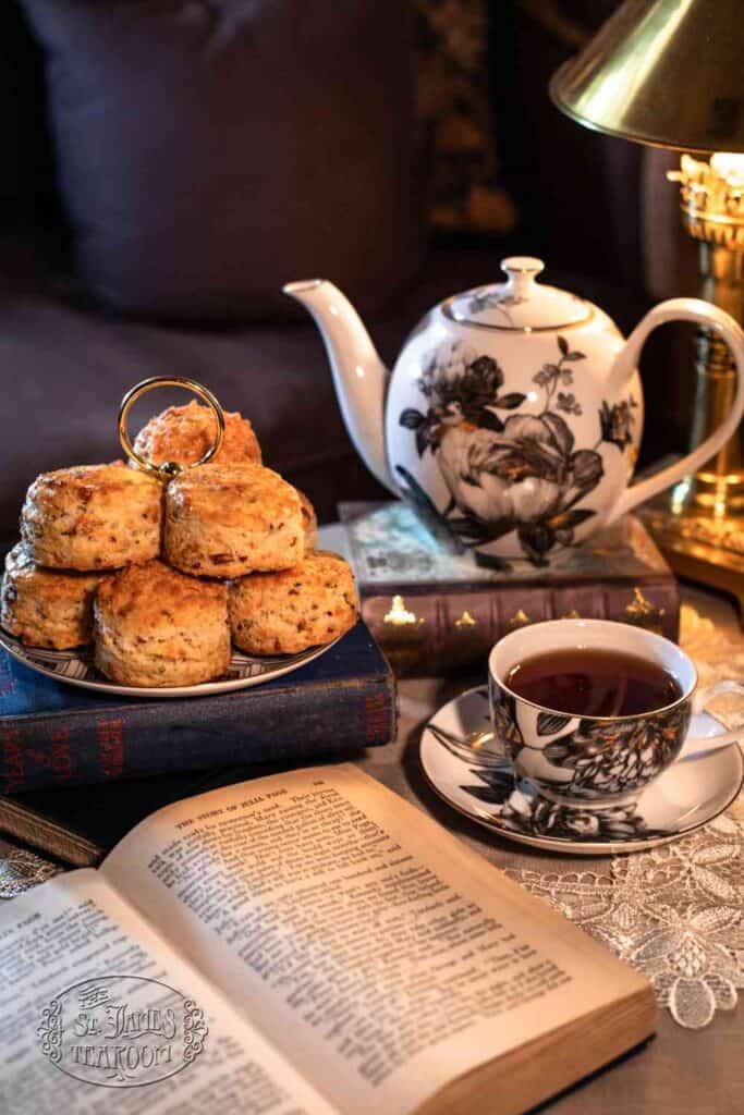 A plate of scones, baked using a classic Buttermilk Scone Recipe, sits alongside a teapot, a teacup with tea, and two books arranged on a lace tablecloth next to a lamp, creating a cozy afternoon tea setting.