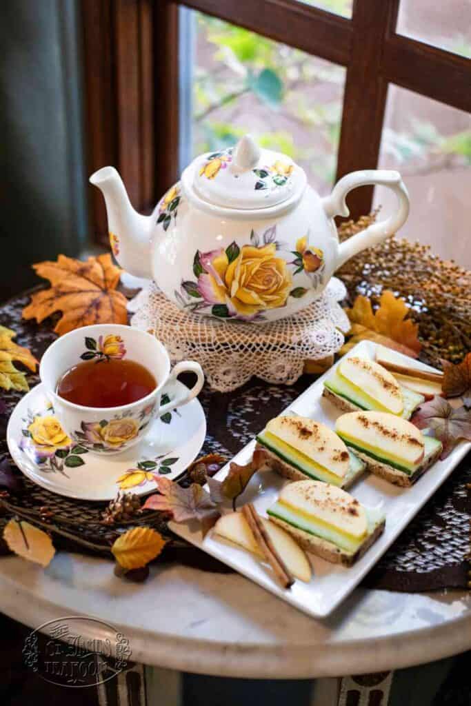 A floral teapot, teacup with tea, and a plate of layered sandwich slices—crafted using the delightful Apple Boursin Tea Sandwich Recipe—sit on a round table decorated with autumn leaves and a lace doily, near a window.