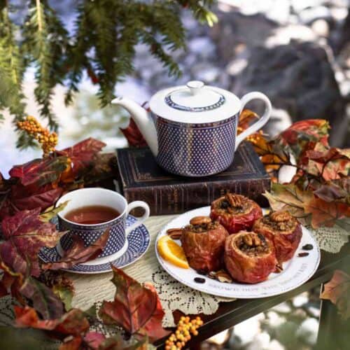 A blue-patterned teapot and cup with tea sit on stacked books beside baked apples on a plate, surrounded by autumn leaves and orange berries, creating a cozy outdoor tea setting that perfectly complements any favorite baked apples recipe.