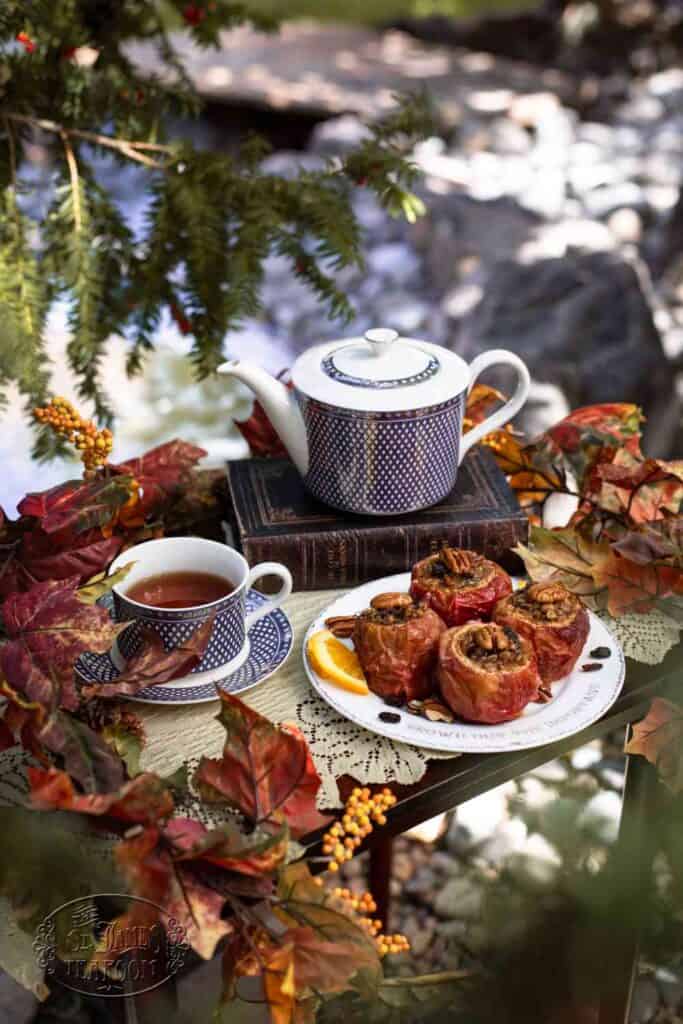 A blue-patterned teapot and cup with tea sit on stacked books beside baked apples on a plate, surrounded by autumn leaves and orange berries, creating a cozy outdoor tea setting that perfectly complements any favorite baked apples recipe.