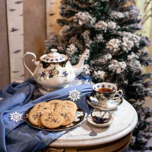 A tea set with a teapot, teacup, and a plate of Earl Grey shortbread cookies—perhaps inspired by your favorite Earl Grey Shortbread Cookies recipe—rests on a small round table, decorated with blue cloth and snowflakes, in front of a snowy, decorated Christmas tree and birch tree backdrop.