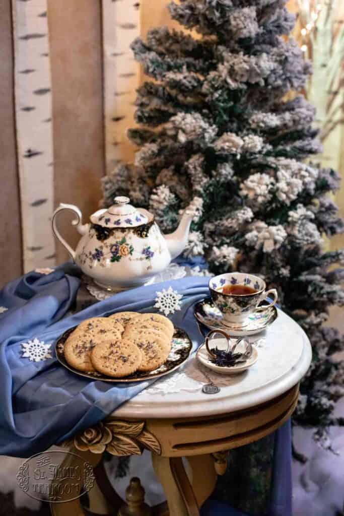 A tea set with a teapot, teacup, and a plate of Earl Grey shortbread cookies—perhaps inspired by your favorite Earl Grey Shortbread Cookies recipe—rests on a small round table, decorated with blue cloth and snowflakes, in front of a snowy, decorated Christmas tree and birch tree backdrop.