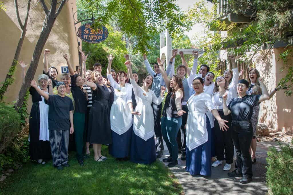 A large group of people, some in costume aprons, smile and cheer outdoors near greenery and a building with a “St. Paul’s Tearoom” sign, appearing joyful and celebratory—perhaps in honor of their recent recognition among Yelp’s Top 100 Restaurants.