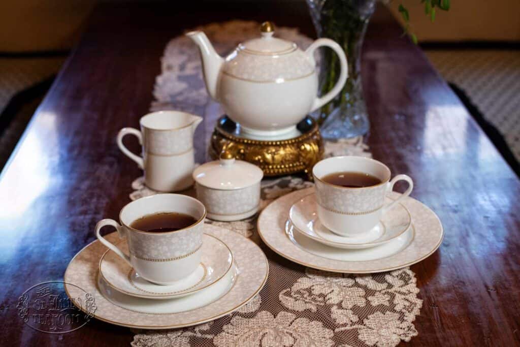 Elegant white and gold tea set with a teapot, sugar bowl, two cups of tea, and two plates on a lace table runner atop a wooden table—a beautiful arrangement that reflects the importance of caring for fine china.