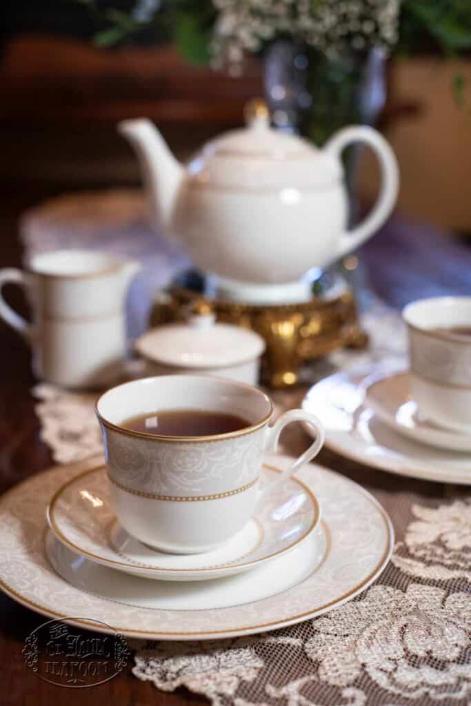 A white porcelain tea set with gold trim, including a teapot, teacups, and saucers, is arranged on a lace tablecloth, with a floral centerpiece blurred in the background—an elegant display that beautifully illustrates the importance of caring for fine china.