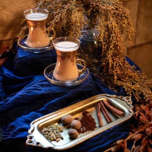 Two ornate glasses of milk tea sit on saucers amid dried flowers. In front, a golden tray holds cinnamon sticks, nutmeg, cardamom pods, and ground spices—essential ingredients for any flavorful iced tea recipe—all beautifully arranged on deep blue fabric with a stone wall background.
