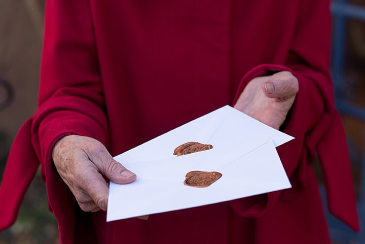 A person in a red coat, reminiscent of someone caring for fine china with delicate attention, holds two white envelopes sealed with brown wax in their hands.