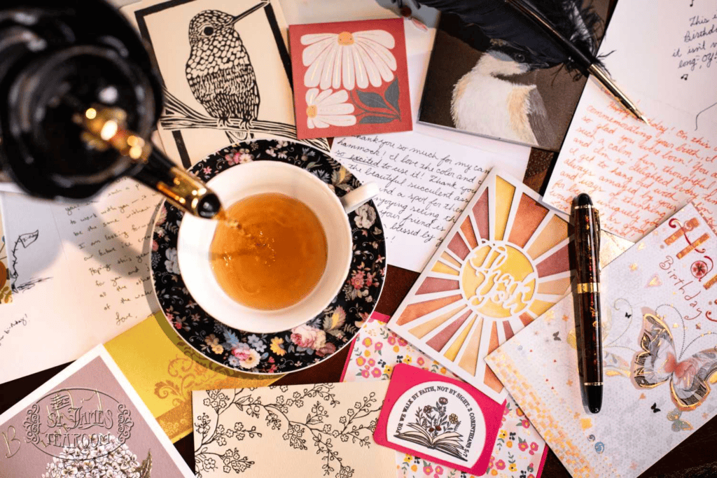A floral teacup with tea being poured sits among handwritten letters, a feather quill, a pen, and colorful illustrated cards featuring flowers, birds, and decorative designs—a charming scene that evokes the timeless ritual of caring for fine china.