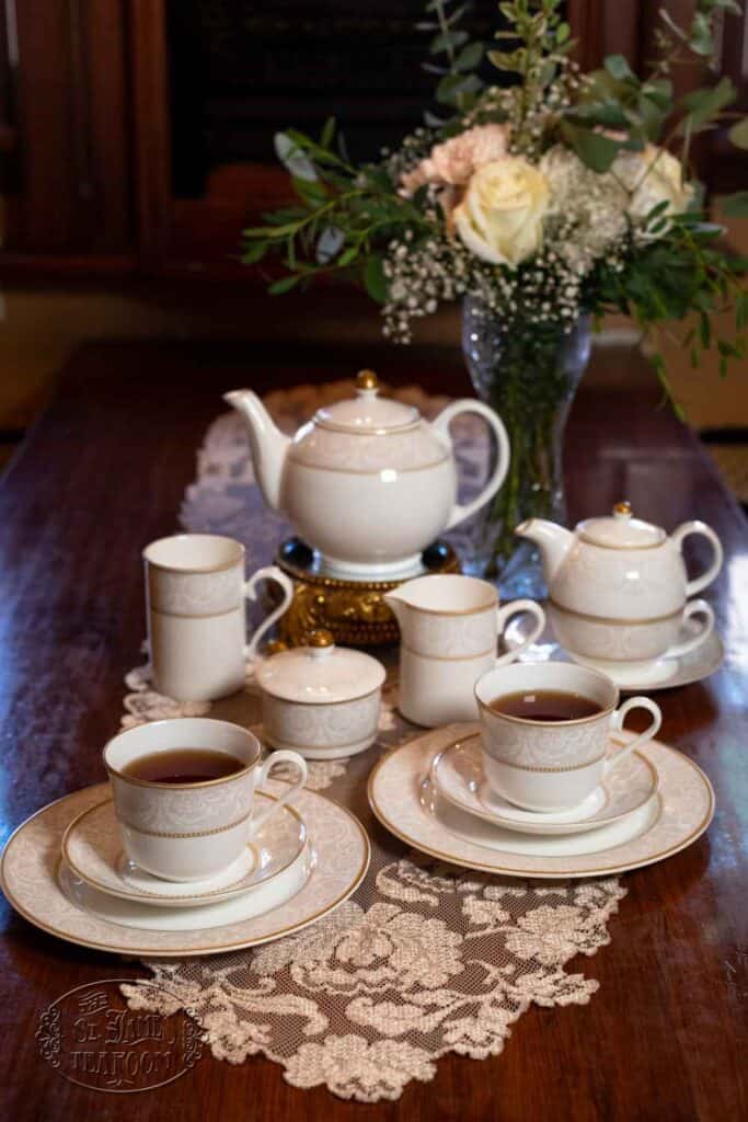 A set of white and gold teacups, saucers, and teapots is arranged on a lace runner atop a wooden table, with a vase of white and cream flowers in the background.