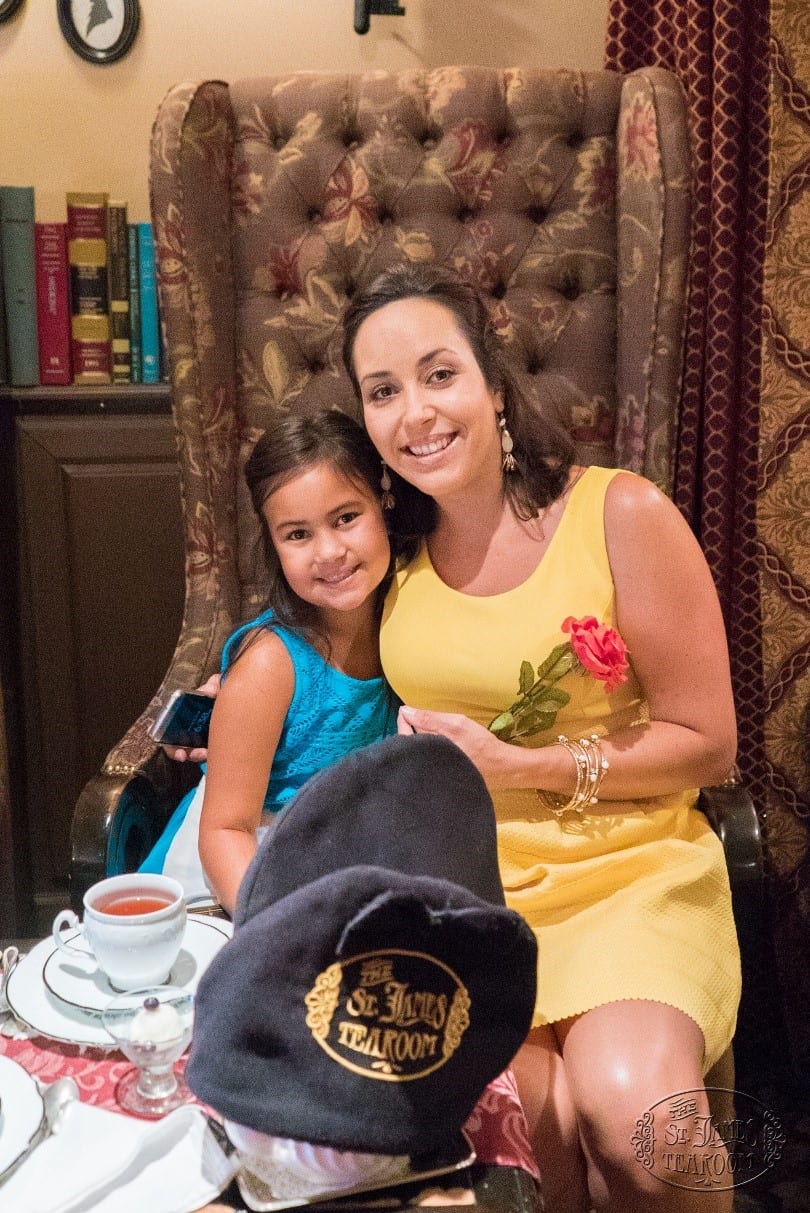 A woman in a yellow dress and a girl in blue smile while sitting together at a tea table, with a cup of tea and a dark hat reading “St. James Tearoom” visible in the foreground.