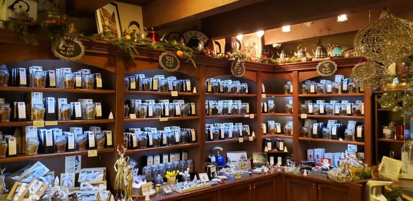 Wooden shelves filled with various packaged teas, tea accessories, and decorative items in a cozy, warmly lit tea shop. Teapots and ornaments are displayed on the top shelf and counter.