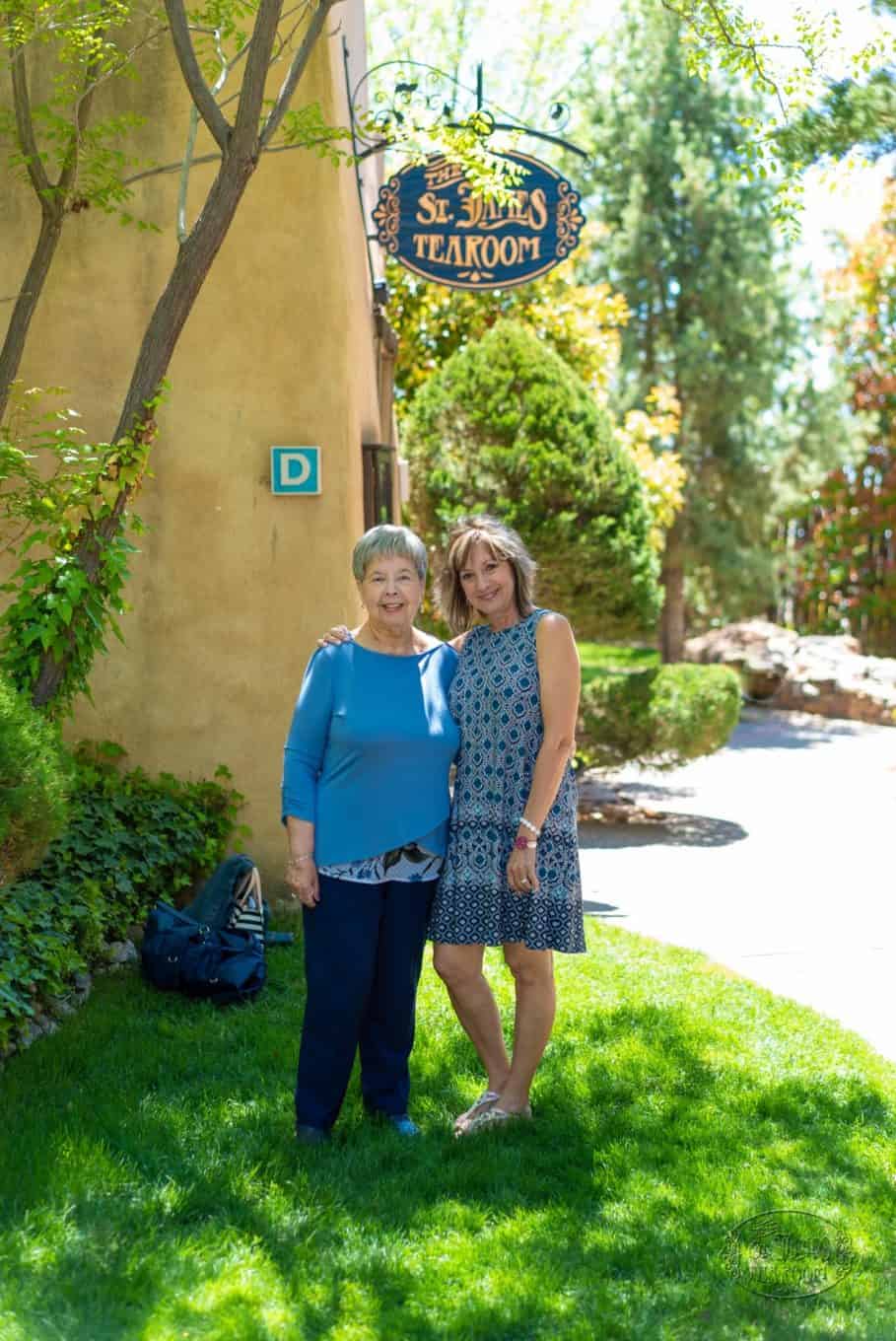 Two women stand smiling on green grass outside, with trees and sunlight around them. A sign above reads “St. James Tearoom.” They look happy and relaxed, enjoying a sunny day together, perhaps about to share a delightful afternoon tea.