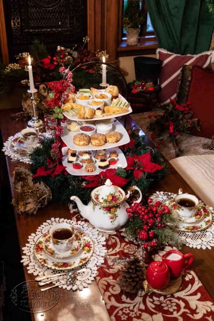 A festive holiday tea setup with a tiered tray of pastries, a floral teapot, teacups, candles, and Christmas decorations, including greenery, pinecones, and red berries, arranged on a lace-covered wooden table.